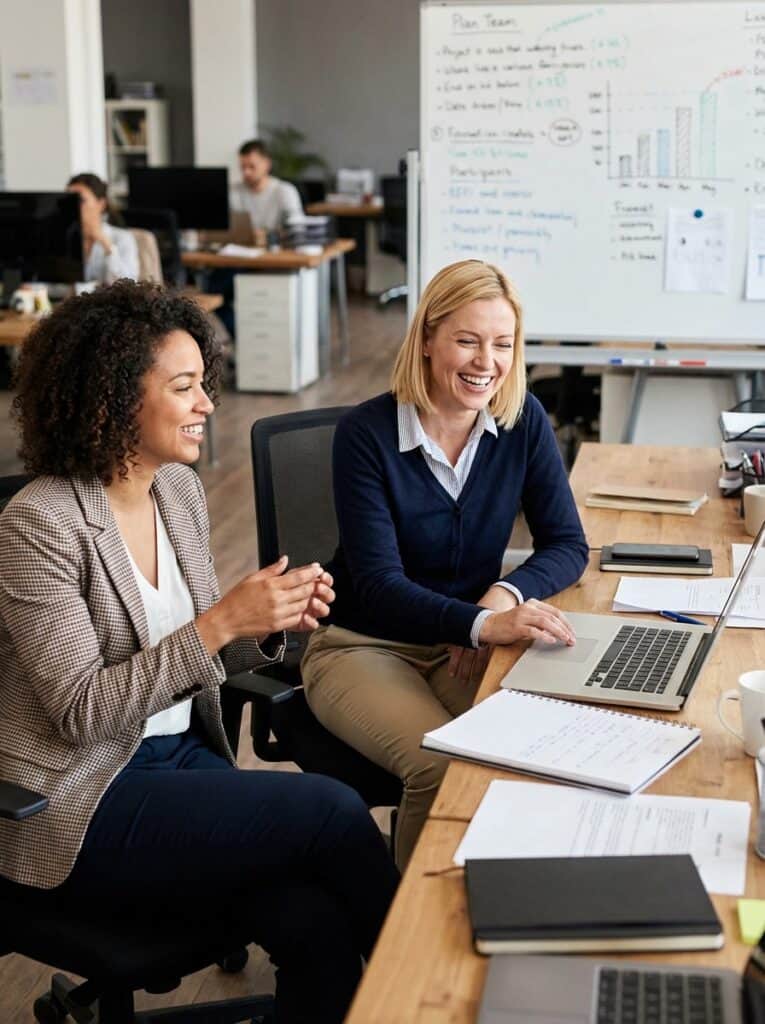 Dos mujeres en una reunión de trabajo en una oficina moderna, compartiendo ideas y sonriendo.